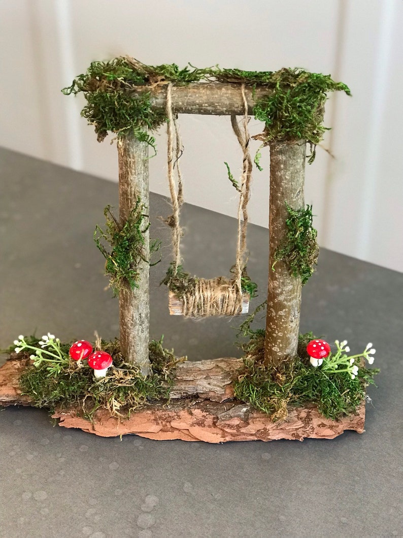 K&ouml;nnte beinhalten: Ein Miniatur-Schaukelgestell aus Holz mit Moos und kleinen roten Pilzen. Die Schaukel besteht aus Schnur und h&auml;ngt an zwei Holzpfosten. Das Schaukelgestell steht auf einem St&uuml;ck Rinde.