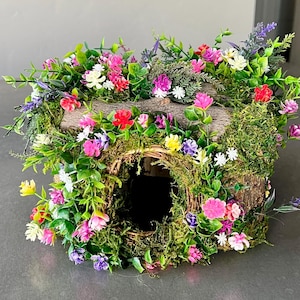 May include: A whimsical, handcrafted birdhouse, adorned with vibrant faux flowers in shades of pink, red, yellow, and white. The birdhouse is constructed from natural wood and moss, creating a charming, rustic aesthetic. The entrance is a circular opening.