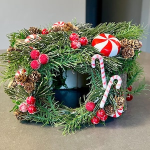 May include: A festive Christmas wreath with green pine needles, red berries, and pine cones. The wreath is decorated with two candy canes and a red and white peppermint candy. A holiday decoration.