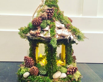 May include: A decorative lantern made of wood and moss, with a glass window and a candle inside. The lantern is decorated with pine cones and birch bark.
