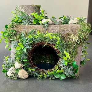 May include: A decorative wooden hamster house with a naturalistic design. The house is made of wood and features a round entrance, surrounded by woven branches. It is adorned with green moss, faux greenery, and small stones, creating a miniature woodland scene.