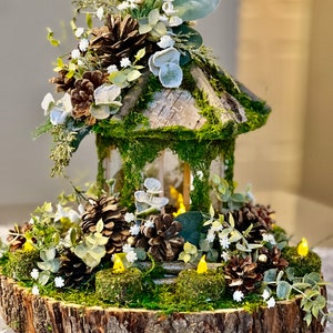 May include: A rustic wooden birdhouse centrepiece decorated with pine cones, moss, and white flowers. The birdhouse is surrounded by a wooden log slice and small LED lights.