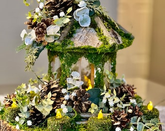 May include: A rustic wooden birdhouse centerpiece decorated with pine cones, moss, and white flowers. The birdhouse is surrounded by a wooden log slice and small LED lights.