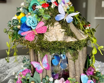 May include: A decorative wooden birdhouse with a moss-covered roof, adorned with colorful paper butterflies, flowers, and miniature mushrooms. The birdhouse features a rustic design with visible wood grain and is surrounded by floral accents.