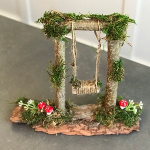 K&ouml;nnte beinhalten: Ein Miniatur-Schaukelgestell aus Holz mit moosbewachsenen Pfosten und einer Schnur-Schaukel. Das Schaukelgestell steht auf einem kleinen St&uuml;ck Rinde mit Moos und zwei roten Pilzen.