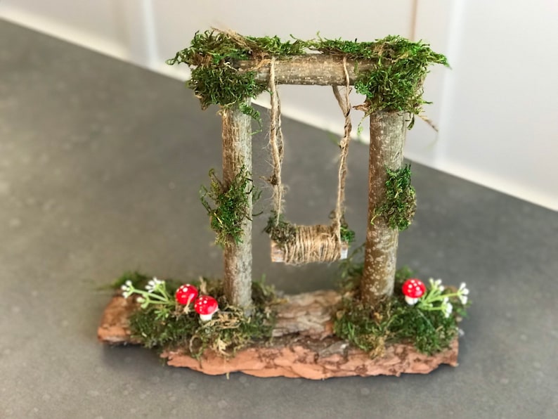 K&ouml;nnte beinhalten: Ein Miniatur-Schaukelgestell aus Holz mit moosbewachsenen Pfosten und einer Schnur-Schaukel. Das Schaukelgestell steht auf einem kleinen St&uuml;ck Rinde mit Moos und zwei roten Pilzen.