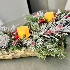 May include: Two rustic log centerpieces featuring artificial evergreen branches with a frosted, snow-dusted finish. Each arrangement includes small brown pinecones, bright red berries, and a single yellow LED pillar candle set into the wood.