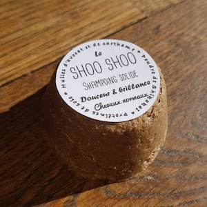 Peut inclure: Un pain de shampooing rond et brun avec une étiquette blanche qui indique "Le SHOO SHOO SHAMPOING SOLIDE Douceur & Brillance Cheveux normaux PROTEINES DE SOIE", un pain de shampooing solide pour cheveux normaux.