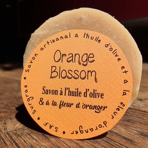 Peut inclure: Un savon rond à la fleur d'oranger avec une étiquette qui indique "Savon artisanal a l'huile d'olive et a la fleur d'oranger S.A.F. Orange Blossom Savon à l'huile d'olive & à la fleur d'oranger"