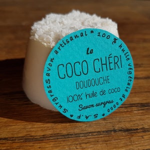 May include: A white, cylindrical soap bar with a topping of shredded coconut. A teal label with French text, including "Le Coco Ch&eacute;ri Doudouche", is attached. The soap sits on a wooden surface.