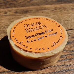 May include: A round bar of orange blossom soap made with olive oil. The soap is wrapped in orange paper with black text that reads "Savon artisanal a l'huile d'olive et a la fleur d'oranger" and "Orange Blossom" in a decorative font. The soap is sitting on a wooden surface.
