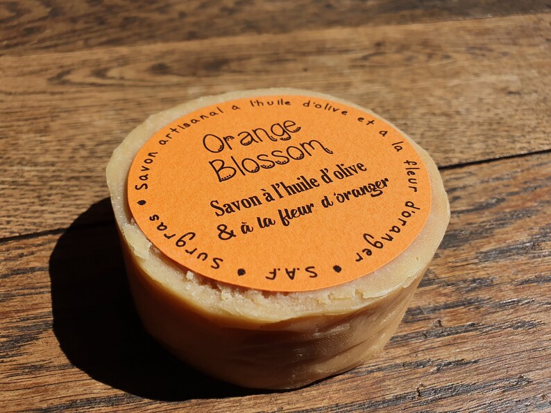 May include: A round bar of orange blossom soap made with olive oil. The soap is wrapped in orange paper with black text that reads "Savon artisanal a l'huile d'olive et a la fleur d'oranger" and "Orange Blossom" in a decorative font. The soap is sitting on a wooden surface.