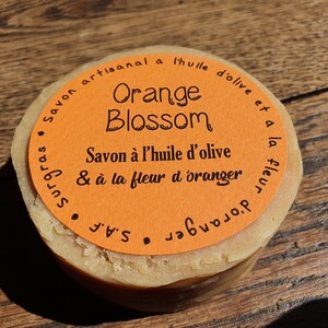 May include: A round orange soap bar with a label that reads "Savon artisanal a l'huile d'olive et a la fleur d'oranger" and "Orange Blossom" in black text.