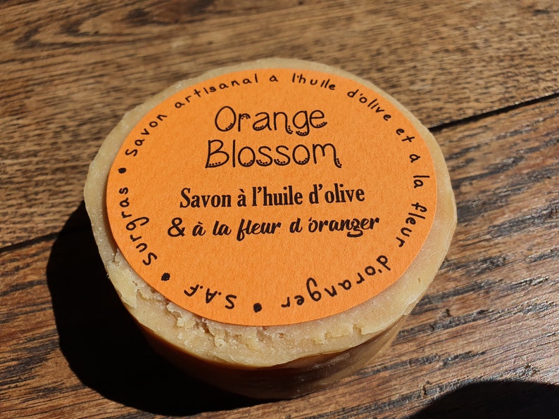 May include: A round orange soap bar with a label that reads "Savon artisanal a l'huile d'olive et a la fleur d'oranger" and "Orange Blossom" in black text.