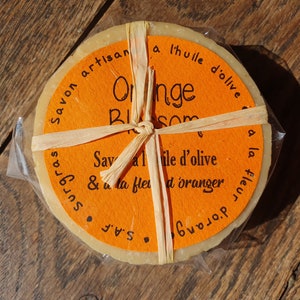 May include: A round bar of orange blossom soap wrapped in brown paper and tied with twine. The label reads "Savon artisan a l'huile d'olive Orange Blossom Savon a l'huile d'olive & a la fleur d'oranger S.A.F. surgras".