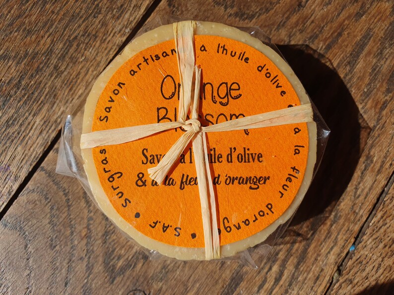 May include: A round bar of orange blossom soap wrapped in brown paper and tied with twine. The label reads "Savon artisan a l'huile d'olive Orange Blossom Savon a l'huile d'olive & a la fleur d'oranger S.A.F. surgras".