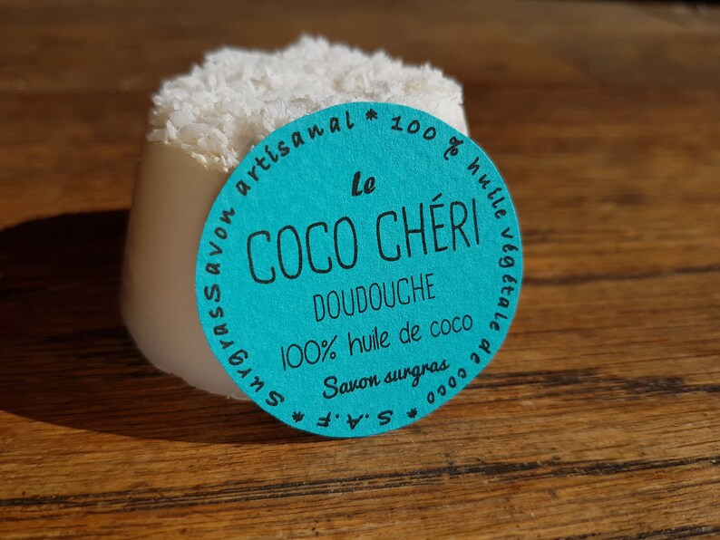 May include: A white, cylindrical soap bar with a topping of shredded coconut. A teal label with French text, including "Le Coco Ch&eacute;ri Doudouche", is attached. The soap sits on a wooden surface.