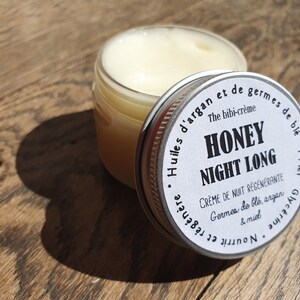 May include: A white jar with a silver lid labeled "Honey Night Long" with the text "The bibi-cr&egrave;me" and "Cr&egrave;me de nuit r&eacute;g&eacute;n&eacute;rante Germes de bl&eacute;, argan & miel Nourrit et r&eacute;g&eacute;n&egrave;re Glycerine Miel" in French. The jar is sitting on a wooden surface.