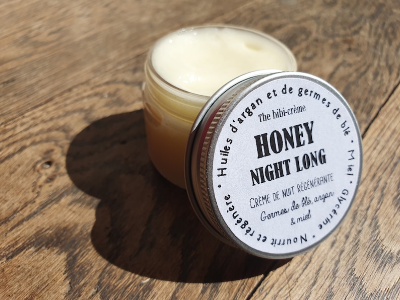 May include: A white jar with a silver lid labeled "Honey Night Long" with the text "The bibi-cr&egrave;me" and "Cr&egrave;me de nuit r&eacute;g&eacute;n&eacute;rante Germes de bl&eacute;, argan & miel Nourrit et r&eacute;g&eacute;n&egrave;re Glycerine Miel" in French. The jar is sitting on a wooden surface.