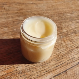 May include: A clear glass jar with a white lid filled with a pale yellow cream.