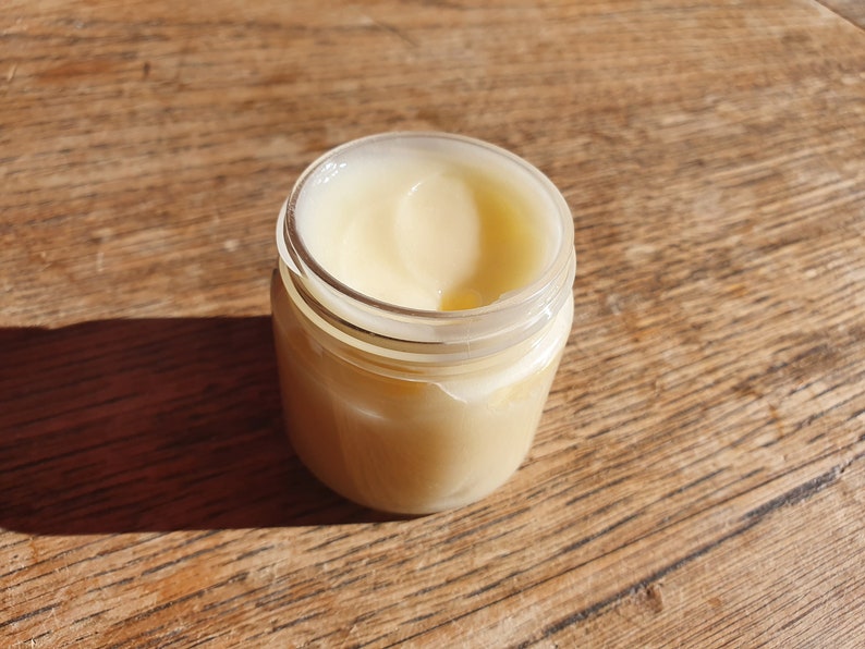 May include: A clear glass jar with a white lid filled with a pale yellow cream.