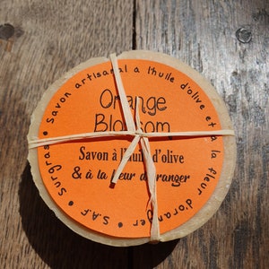 May include: Two round bars of orange blossom soap wrapped in twine. The soap is made with olive oil and is artisanally crafted. The label reads "Savon artisanal a l'huile d'olive et surgras, Orange Blossom, Savon a l'huile d'olive & a la fleur d'oranger, S.A. "