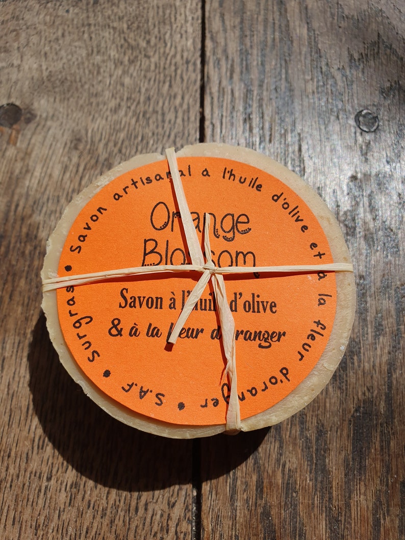 May include: Two round bars of orange blossom soap wrapped in twine. The soap is made with olive oil and is artisanally crafted. The label reads "Savon artisanal a l'huile d'olive et surgras, Orange Blossom, Savon a l'huile d'olive & a la fleur d'oranger, S.A. "