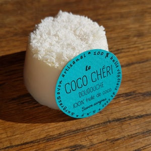 May include: A white soap bar with a shredded coconut topping. A teal label with French text, including "Le Coco Ch&eacute;ri Doudouche", is attached. The soap sits on a wooden surface. The soap is approximately 7 cm in diameter.