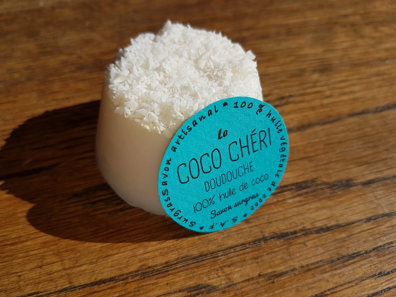 May include: A white soap bar with a shredded coconut topping. A teal label with French text, including "Le Coco Ch&eacute;ri Doudouche", is attached. The soap sits on a wooden surface. The soap is approximately 7 cm in diameter.