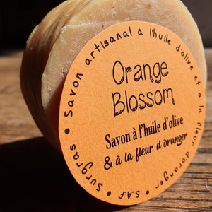 May include: A round orange soap bar with a label that reads "Savon artisanal a l'huile d'olive et a la fleur d'oranger" and "Orange Blossom".