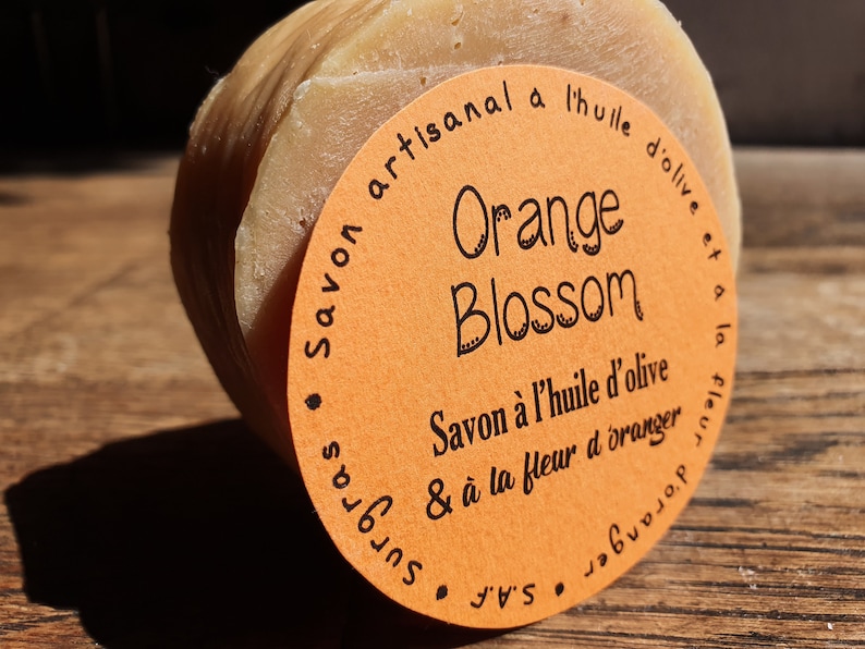 May include: A round orange soap bar with a label that reads "Savon artisanal a l'huile d'olive et a la fleur d'oranger" and "Orange Blossom".