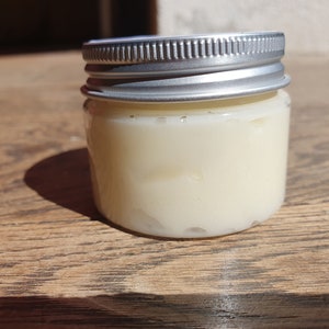 May include: A small glass jar with a silver lid filled with a white cream. The jar is sitting on a wooden surface.