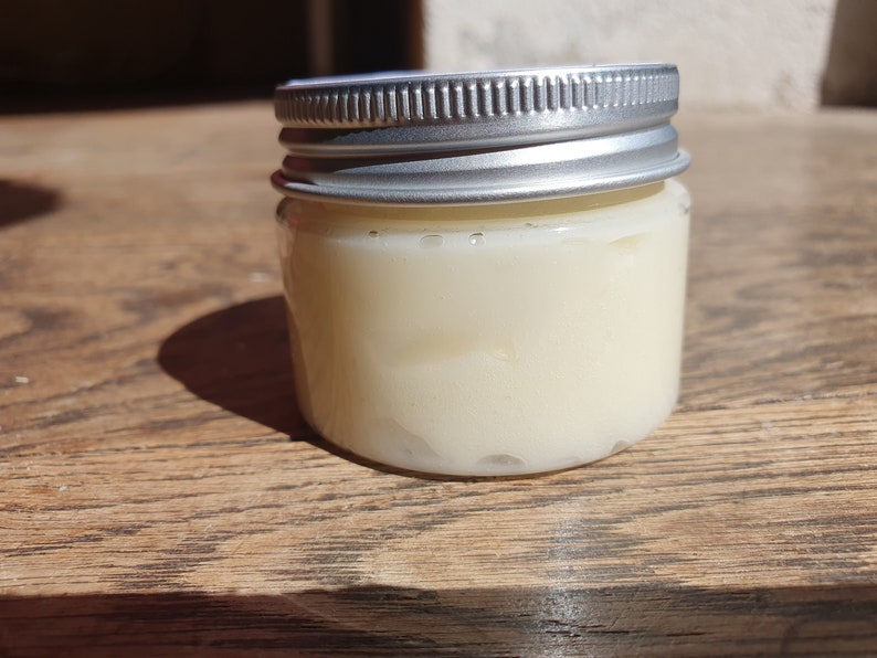 May include: A small glass jar with a silver lid filled with a white cream. The jar is sitting on a wooden surface.
