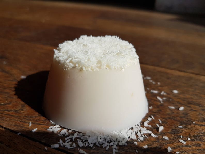 May include: A white, dome-shaped dessert, possibly a panna cotta, topped with shredded coconut. The dessert is on a dark wooden surface, with scattered coconut flakes around it.