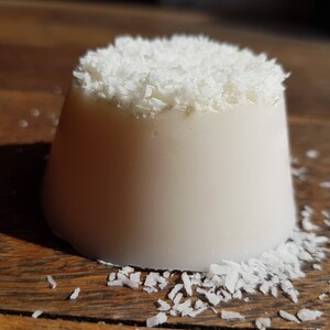 May include: A white, cylindrical dessert, topped with shredded coconut, rests on a wooden surface. The dessert has a smooth, creamy appearance, with coconut flakes on top and scattered around the base.