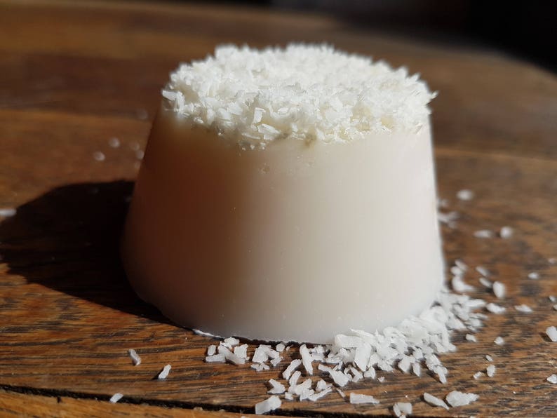 May include: A white, cylindrical dessert, topped with shredded coconut, rests on a wooden surface. The dessert has a smooth, creamy appearance, with coconut flakes on top and scattered around the base.