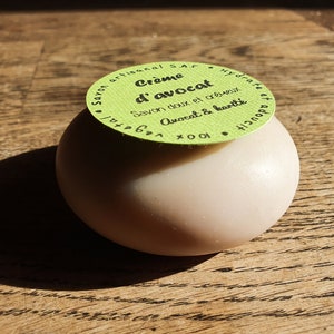May include: A green and white round label with the text "Cr&egrave;me d'avocat Savon doux et cr&eacute;meux Avocat & karit&eacute; 100% v&eacute;g&eacute;tal Hydrate et adoucit" on top of a round, light brown soap bar.