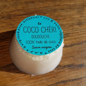 May include: A white, egg-shaped soap with a turquoise label that reads "Le Coco Ch&eacute;ri Doudouche 100% huile de coco Savon surgras". The soap has a dusting of white flakes around the top and sits on a wooden surface.