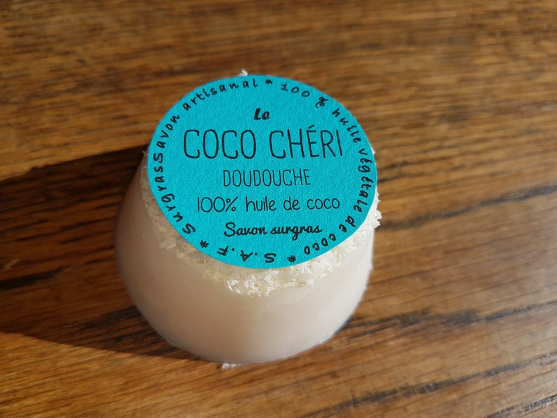 May include: A white, egg-shaped soap with a turquoise label that reads "Le Coco Ch&eacute;ri Doudouche 100% huile de coco Savon surgras". The soap has a dusting of white flakes around the top and sits on a wooden surface.