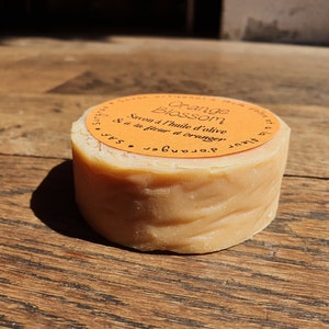 May include: A round bar of orange blossom soap made with olive oil. The soap is wrapped in a bright orange label with the text "Orange Blossom Savon &agrave; l'huile d'olive &agrave; la fleur d'oranger".