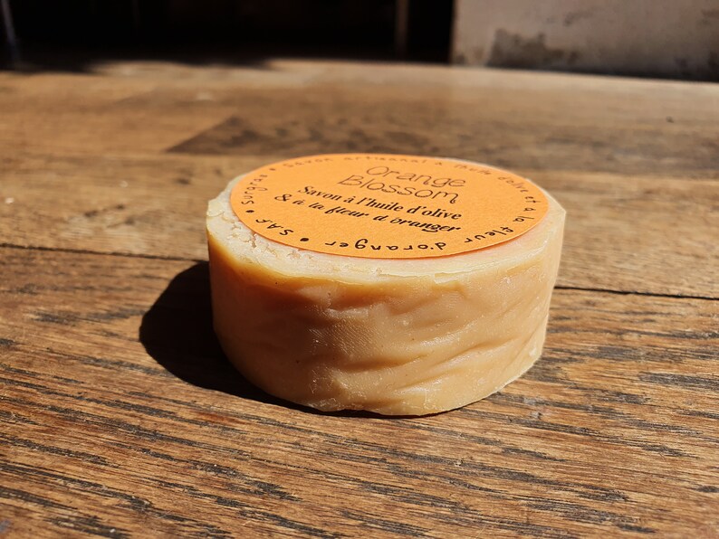 May include: A round bar of orange blossom soap made with olive oil. The soap is wrapped in a bright orange label with the text "Orange Blossom Savon &agrave; l'huile d'olive &agrave; la fleur d'oranger".