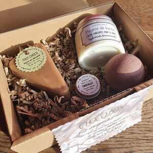 Puede incluir: Una caja de regalo de cart&oacute;n marr&oacute;n llena de productos de ba&ntilde;o con tem&aacute;tica de chocolate. La caja contiene un jab&oacute;n con aroma a chocolate, una bomba de ba&ntilde;o de chocolate, un peque&ntilde;o tarro de manteca corporal de chocolate y una etiqueta que dice "CHOCO-BOX Pour une peau &agrave; croquer".
