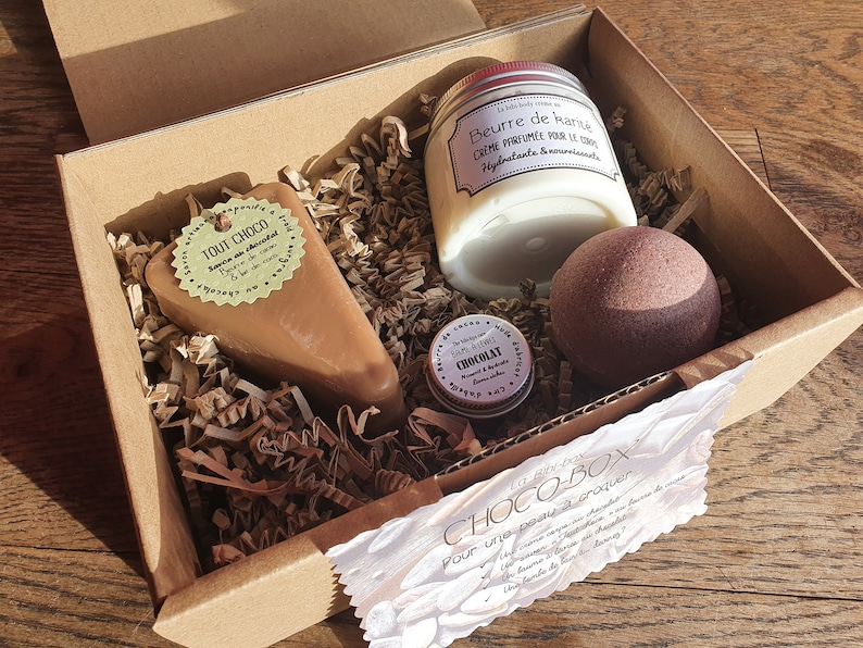 Puede incluir: Una caja de regalo de cart&oacute;n marr&oacute;n llena de productos de ba&ntilde;o con tem&aacute;tica de chocolate. La caja contiene un jab&oacute;n con aroma a chocolate, una bomba de ba&ntilde;o de chocolate, un peque&ntilde;o tarro de manteca corporal de chocolate y una etiqueta que dice "CHOCO-BOX Pour une peau &agrave; croquer".