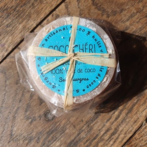 May include: A circular soap bar, light brown in colour, wrapped in clear plastic and tied with raffia. The soap features a blue label with the text "Coco Ch&eacute;ri" and "100% huile de coco". It rests on a wooden surface.