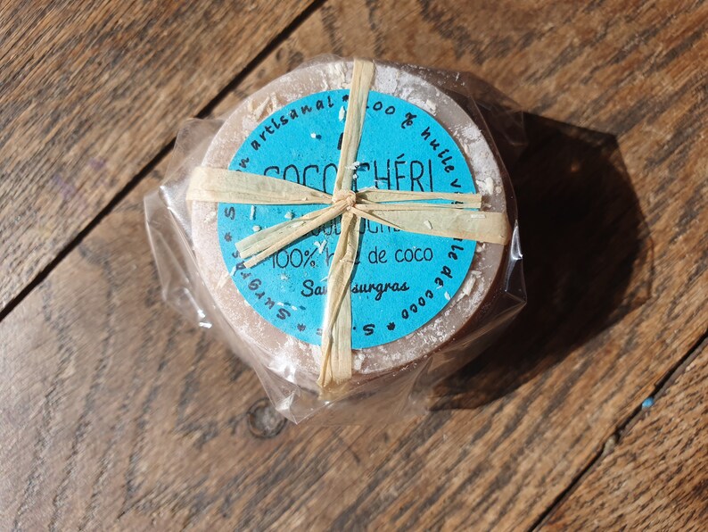 May include: A circular soap bar, light brown in colour, wrapped in clear plastic and tied with raffia. The soap features a blue label with the text "Coco Ch&eacute;ri" and "100% huile de coco". It rests on a wooden surface.