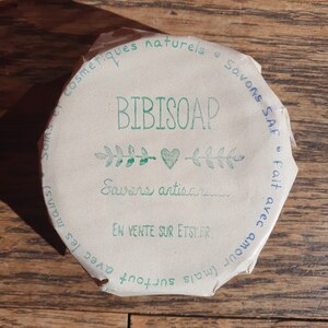 May include: A close-up of a round, brown paper label with green text that reads "BIBISOAP" and "Savons artisanaux" with a green heart and leaves. The label also includes the text "Cosmetiques naturels", "Savons SAF", "En vente sur Etsy.fr", "Avec amour (mais surtout avec les mains)".