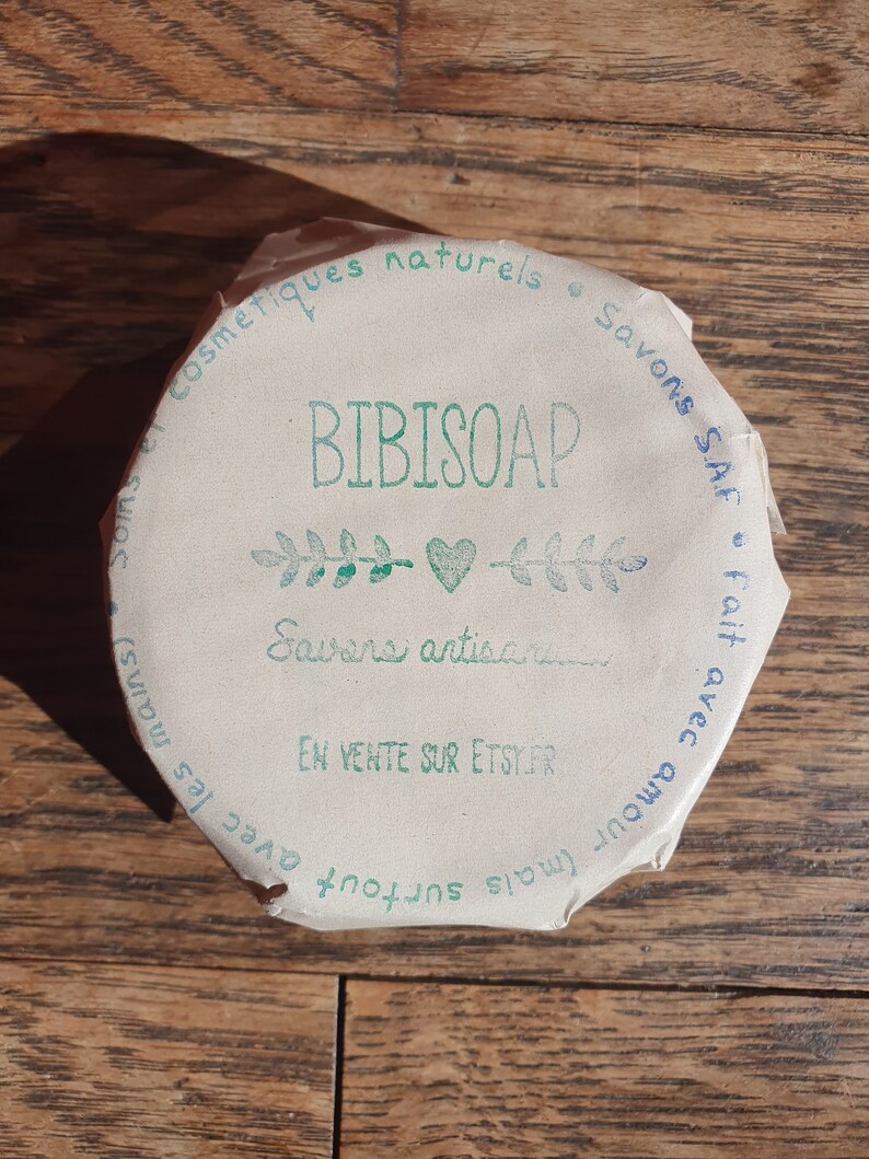 May include: A close-up of a round, brown paper label with green text that reads "BIBISOAP" and "Savons artisanaux" with a green heart and leaves. The label also includes the text "Cosmetiques naturels", "Savons SAF", "En vente sur Etsy.fr", "Avec amour (mais surtout avec les mains)".