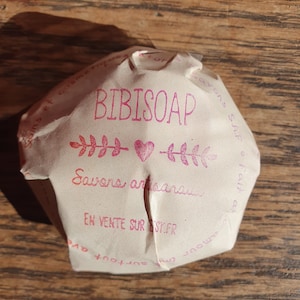 May include: A white paper wrapper with pink text that reads "BIBISOAP" and "Savons artisanaux" with a pink heart symbol between two sets of pink leaf symbols. The text "EN VENTE SUR ST.FR" is also printed in pink.
