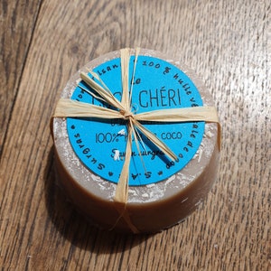May include: A circular, light brown soap bar with a blue label and text, tied with raffia. The label says "le CH&Eacute;RI" and "100% huile v&eacute;". The soap is presented on a wooden surface.