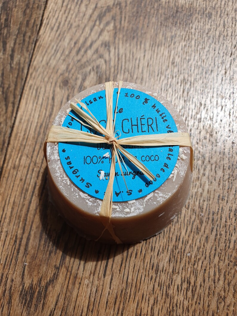 May include: A circular, light brown soap bar with a blue label and text, tied with raffia. The label says "le CH&Eacute;RI" and "100% huile v&eacute;". The soap is presented on a wooden surface.